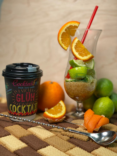 Abbildung des Glüh-Cocktails: Cocktailglas mit Rohrzucker, Limetten- und Orangenscheiben. Daneben ein To-Go Becher mit dem Cocktailooo Logo und der Aufschrift 