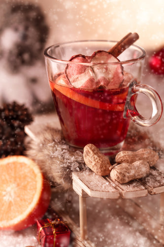 Abbildung des Glüh-Cocktails: Glas-Tasse mit klar-roter Flüssigkeit. Daneben Äpfel und Zimtrollen und Erdnüsse. Schneeflocken fallen herab.