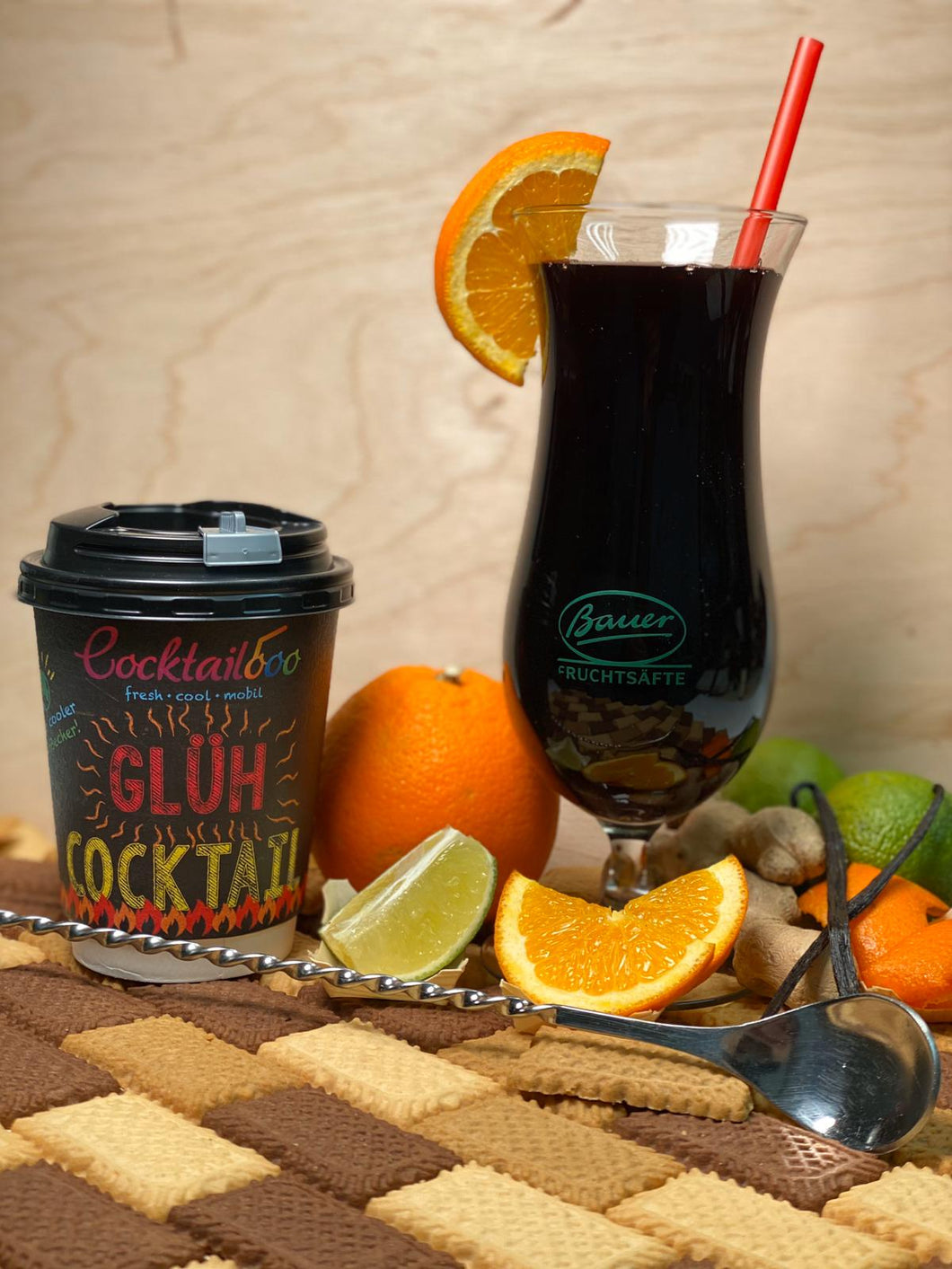 Abbildung des Glüh-Cocktails: Cocktailglas mit dunkelroter Flüssigkeit. Daneben ein To-Go Becher mit dem Cocktailooo Logo und der Aufschrift 